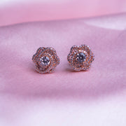 A pair of rose gold-colored evening primrose stud earrings displayed against a pink background.